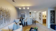 Maple Brook Apartments presents a thoughtfully designed single-floor residence that seamlessly blends comfort and contemporary style. The open-concept living area is anchored by plush carpeting and soft, neutral-toned walls that create a warm and inviting atmosphere. Track lighting overhead provides ample illumination, highlighting the space's clean lines and modern aesthetic.

The living area flows naturally into a casual dining nook, complete with a bistro-style table and seating — perfect for intimate meals or casual gatherings. Adjacent to the dining area, the kitchen features a peninsula-style breakfast bar with stylish bar stools, offering additional seating and a natural gathering point. Warm wood cabinetry and light countertops lend a timeless look to the kitchen.

Decorative wall accents, including a framed cityscape artwork and a set of sunburst mirrors, add personality and visual interest throughout the common areas. A hallway leads to additional rooms, with a glimpse of a bathroom vanity suggesting well-appointed secondary spaces. The overall design balances functional living with tasteful décor, making Maple Brook Apartments an appealing and comfortable place to call home.