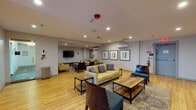 This single-floor communal lounge is thoughtfully designed to encourage comfort and social interaction. The space is anchored by light wood flooring that runs throughout, creating a warm and cohesive aesthetic. Recessed lighting illuminates the room evenly, complementing the neutral gray and soft yellow walls that lend a clean, contemporary feel.

The lounge is divided into distinct seating zones, each furnished with a mix of cream and yellow upholstered sofas and chairs accented with blue and gray throw pillows. Dark wood and metal-frame coffee tables are positioned throughout, offering practical surfaces for each seating group. Teal-cushioned benches and dark wood slatted accent chairs add visual contrast and variety.

A wall-mounted flat-screen television, positioned above a dark wood media console, serves as a focal point for one of the primary seating areas. A second TV is also mounted on an opposing wall, ensuring multiple viewing options across the space. Framed abstract artwork in muted tones decorates the walls, adding a polished, curated touch.

A wall-mounted mini-split air conditioning unit provides climate control, and the space is equipped with standard safety features including a fire suppression system and emergency exit signage. A large window dressed with vertical blinds allows natural light to filter in, enhancing the overall ambiance of this inviting, multifunctional gathering space.