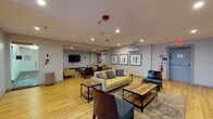 This single-floor communal lounge is thoughtfully designed to encourage comfort and social interaction. The space is anchored by light wood flooring that runs throughout, creating a warm and cohesive aesthetic. Recessed lighting illuminates the room evenly, complementing the neutral gray and soft yellow walls that lend a clean, contemporary feel.

The lounge is divided into distinct seating zones, each furnished with a mix of cream and yellow upholstered sofas and chairs accented with blue and gray throw pillows. Dark wood and metal-frame coffee tables are positioned throughout, offering practical surfaces for each seating group. Teal-cushioned benches and dark wood slatted accent chairs add visual contrast and variety.

A wall-mounted flat-screen television, positioned above a dark wood media console, serves as a focal point for one of the primary seating areas. A second TV is also mounted on an opposing wall, ensuring multiple viewing options across the space. Framed abstract artwork in muted tones decorates the walls, adding a polished, curated touch.

A wall-mounted mini-split air conditioning unit provides climate control, and the space is equipped with standard safety features including a fire suppression system and emergency exit signage. A large window dressed with vertical blinds allows natural light to filter in, enhancing the overall ambiance of this inviting, multifunctional gathering space.