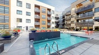 This expansive outdoor amenity courtyard is set within a contemporary multi-story building featuring a modern architectural design with mixed exterior cladding in neutral tones, warm wood accents, and glass-railed balconies on all surrounding floors. The centerpiece of the space is a large, resort-style swimming pool with vivid turquoise water, flanked by rows of bold orange lounge chairs and sleek gray chaise lounges that create a vibrant, energetic atmosphere. A shallow wading ledge runs alongside the pool, providing additional relaxation space. Adjacent to the pool, a built-in fire pit lounge area offers generous seating with deep cushioned benches adorned with bright orange and pink pillows, arranged around a long linear gas fire feature set in stone tile. Colorful red folding tables and chairs are scattered throughout for casual outdoor dining. A covered outdoor kitchen station houses a stainless steel grill and storage drawers beneath a slatted pergola strung with globe lights. Lush ornamental grasses and planters are integrated throughout, softening the hardscape. The courtyard opens to sweeping views of a mountain range and surrounding urban skyline, completing the outdoor experience.