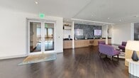 Located at 525 NE 7th St in Fort Lauderdale, Florida, this stylish amenity lounge spans a generously sized single floor designed to blend comfort, functionality, and bold contemporary aesthetics. The main lounge area immediately captures attention with its large floor-to-ceiling windows that flood the space with natural light and offer open views of swaying palms and the South Florida skyline. Dark hardwood-style flooring runs throughout, grounding the vibrant, eclectic furnishings that include plush velvet seating in deep purples, pinks, and navy blues.

A striking oversized mural of a cat wearing sunglasses serves as a bold focal point, complementing the curated collection of colorful abstract artwork displayed along the corridors. The refreshment bar area features warm wood cabinetry, dark stone countertops, and a dramatic textured stone accent wall with integrated display screens — ideal for hosting community events and announcements.

A dedicated conference or meeting room is enclosed with glass walls, furnished with a round table and modern transparent chairs, offering a private yet visually connected workspace. The connecting hallway features vibrant patchwork carpeting in oceanic blues and earth tones, adding a creative transition between zones. Recessed lighting throughout ensures a bright, welcoming ambiance at all times, making this a dynamic and engaging communal destination.