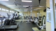 This fitness center is a comprehensively outfitted exercise facility designed to accommodate a variety of workout routines. The space is generously sized, providing ample room for movement between multiple stations. The flooring consists of durable dark rubber matting, which offers both safety and comfort during exercise.

The room is furnished with an extensive selection of strength-training equipment, including cable machines, weight benches, and multi-function resistance stations arranged throughout the floor. Along one side of the space, a row of cardiovascular machines — including ellipticals and treadmills — lines the wall, offering dedicated space for aerobic workouts.

Natural light filters in through windows along the perimeter, complemented by recessed fluorescent panel lighting across the ceiling to ensure consistent, bright illumination throughout the room. Multiple ceiling fans are installed overhead, promoting air circulation and maintaining a comfortable workout environment. Large wall mirrors enhance the sense of space while allowing users to monitor their form. The warm yellow-toned walls contribute to an inviting, energetic atmosphere well-suited for fitness activity.