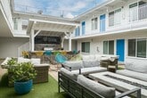 Located at 60 15th St in the heart of Hermosa Beach, California, this inviting property centers around a beautifully designed open-air courtyard that serves as the social hub of the community. The architecture features a crisp white exterior with bold blue accent doors and white railings along the upper-level balconies, evoking a classic Southern California coastal aesthetic.

The expansive courtyard is thoughtfully arranged with multiple seating zones, including plush sectional sofas, wicker lounge sets, and colorful accent chairs positioned atop patches of artificial turf and stone tile pathways. String lights crisscross overhead, creating a warm and festive ambiance ideal for evening gatherings. A wall-mounted flat-screen television anchored to a wood-panel accent wall serves as a focal point for one of the lounge areas, while a covered entertainment zone at the far end features a built-in grill and bar-height seating surrounded by vibrant yellow and teal chairs.

A dedicated storage area beneath the upper balcony provides convenient space for bicycles and surfboards, catering perfectly to the active beach lifestyle that defines the neighborhood. Lush potted greenery adds natural warmth throughout the courtyard. The overall atmosphere is relaxed yet lively, blending resort-style outdoor living with the laid-back charm of one of Los Angeles County's most beloved beachside communities.
