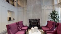 This refined hospitality space makes an immediate impression with its dramatic, double-height lobby ceiling and stately architectural detailing. White wainscoting and paneled walls frame the main lounge area, where a stunning floor-to-ceiling marble fireplace surround serves as the focal centerpiece. Plush, jewel-toned wingback chairs are arranged around a marble-topped coffee table, creating an inviting seating area perfect for relaxed conversation or quiet reflection. A grand curved staircase with dark-painted turned balusters and a polished metal newel post adds a touch of classic grandeur, its treads dressed in a charcoal-toned runner. Adjacent to the lobby, a business center offers a compact yet fully functional workspace. Built-in warm wood-panel cabinetry lines the walls, complemented by an L-shaped white countertop workstation equipped with desktop computers, a printer, and wall-mounted monitor. The space is accented by a bold, graphic black-and-white mural that energizes the room with a modern artistic contrast. Wood-look tile flooring and recessed lighting complete the contemporary-meets-classic aesthetic throughout. Patterned carpeting visible through glass partitions adds further visual interest to the connecting corridors, tying together the space's layered design sensibility.