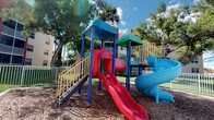 This outdoor amenity space offers a well-appointed environment designed for recreation and relaxation. The playground area is a standout feature, showcasing a multi-structure play system with vivid primary colors. It includes a red open slide, a red tunnel slide, a blue spiral slide, and multiple platform levels connected by stairways with yellow handrails. The structures are supported by bold blue and green posts, topped with teal and green canopy roofs. The ground beneath is layered with wood chip mulch, providing a safe, impact-absorbing surface. The playground is enclosed by a white metal fence, ensuring a secure play environment. Mature trees surround the area, offering natural shade and a lush tropical backdrop.

Adjacent to the playground, a serene lakefront area adds a tranquil counterpoint to the active play space. A white metal fence lines the water's edge, framing expansive views of the lake, open sky, and neighboring buildings across the water. The grassy lakeside lawn provides a pleasant open area to enjoy the scenic waterfront setting. Together, these amenities create a well-rounded outdoor space suited for both active play and peaceful leisure.