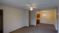 This single-floor residence offers a practical and comfortable layout designed for everyday living. The main living area is generously sized, finished with neutral wall-to-wall carpeting and bright white walls that create a clean, open atmosphere. A ceiling fan with an integrated light fixture provides both comfort and illumination, while a decorative chandelier marks the transition into the adjacent dining area, adding a touch of elegance to the space.

The open-concept flow between the living and dining areas allows for versatile furniture arrangements and easy movement throughout. The kitchen is positioned just beyond, featuring warm medium-toned wood cabinetry that adds a classic touch. A countertop and base cabinets provide ample storage and workspace, making the kitchen both functional and inviting.

Throughout the space, consistent carpeting and neutral tones create a cohesive visual experience. Recessed ventilation and thoughtful lighting placements contribute to a well-maintained interior environment. The overall ambiance is understated and move-in ready, suited to a variety of lifestyles. The single-level layout ensures ease of accessibility and a seamless connection between all living spaces.