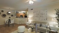 This single-story residence showcases an open-concept layout that seamlessly connects the living area and kitchen, creating a spacious, airy feel throughout. The living room is finished with warm neutral tones and features elegant crown molding along the ceiling, adding a refined architectural touch. A ceiling fan anchors the space, contributing both style and function. The room comfortably accommodates a plush sofa adorned with decorative pillows, a glass coffee table, and a rounded ottoman, all arranged atop a light area rug over hardwood-style flooring. Contemporary wall art adds a modern accent to the neutral palette. The adjoining kitchen is positioned on a raised counter-height platform, offering a breakfast bar with seating for casual dining. It features warm wood cabinetry, dark countertops, and black appliances, including a built-in microwave. Overhead lighting illuminates the kitchen workspace effectively. The open sightlines between the kitchen and living area enhance the sense of connectivity and spaciousness throughout the home, making it well-suited for both everyday living and entertaining.