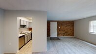 Located at 203–247 NC-54 W in Carrboro, NC, this single-floor unit offers a well-organized layout with distinct living, sleeping, and kitchen areas. The living room makes an immediate impression with a full exposed brick accent wall framing the entryway, complemented by plush carpet flooring and a window fitted with blinds that allows natural light to fill the space. The kitchen is a functional galley-style design, featuring grey shaker-style cabinetry, light stone-look countertops, a double stainless steel sink, and stainless steel appliances including a gas range, dishwasher, and refrigerator. A brick tile backsplash and light hardwood-style flooring complete the kitchen's clean, modern aesthetic. The bedrooms are carpeted and offer a quiet, neutral atmosphere; the primary bedroom includes a ceiling fan with light fixture, a window with blinds, and two separate closet doors. An additional room with carpet extends the flexible living space. The bathroom features a full tub and shower combination with white subway tile surround, brushed nickel grab bar, and classic mosaic tile flooring. Throughout the unit, neutral wall tones and consistent finishes create a cohesive and move-in-ready environment.