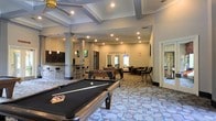 This single-floor amenity space is thoughtfully designed as a multifunctional recreation and social hub. The open-plan layout is anchored by a full-size billiards table positioned prominently in the foreground, complemented by a foosball table nearby, making it a centerpiece for game-day gatherings and casual entertainment. The room features an elevated tray ceiling with decorative exposed beams and recessed lighting, adding architectural interest and a sense of volume to the space. Structural columns with decorative detailing define zones within the open floor plan without disrupting the flow of the room. A wet bar area with dark cabinetry, pendant lighting, and bar-height seating lines one side of the space, providing a convenient refreshment station. Patterned carpeting in muted, earthy tones covers the floor, adding texture and visual interest throughout. Toward the rear of the room, additional seating and gaming areas are visible, along with wall-mounted televisions for viewing. Natural light filters in through windows and glass doors at the far end, brightening the space. The overall ambiance is lively yet comfortable, well-suited for community socializing and recreational activities.