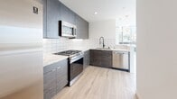 This contemporary single-floor unit showcases a clean, modern design throughout. The living area is a standout, featuring a dramatic curved wall of floor-to-ceiling windows that flood the space with natural light and provide open urban views, complemented by a balcony access door framed by sheer white curtains. Light wood-tone flooring runs consistently throughout, enhancing the airy, cohesive aesthetic.

The kitchen is well-appointed with dark wood-grain cabinetry extending to the ceiling, quartz countertops with subtle veining, a geometric white tile backsplash, and a full suite of stainless steel appliances including a gas range, microwave, and dishwasher. Pendant lighting and a large window add brightness to the workspace.

Bedrooms feature large windows with neighborhood views, modern gold-accented ceiling fans with recessed lighting, and frosted glass sliding closet doors in at least one room. The primary bathroom offers a sleek dark wood vanity, an LED-backlit mirror, white tile flooring, and a tub/shower combination with striking marble-look porcelain tile surround and frameless glass slider. A secondary half bath provides additional convenience with a matching minimalist design.