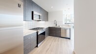 This contemporary single-floor unit showcases a clean, modern design throughout. The living area is a standout, featuring a dramatic curved wall of floor-to-ceiling windows that flood the space with natural light and provide open urban views, complemented by a balcony access door framed by sheer white curtains. Light wood-tone flooring runs consistently throughout, enhancing the airy, cohesive aesthetic.

The kitchen is well-appointed with dark wood-grain cabinetry extending to the ceiling, quartz countertops with subtle veining, a geometric white tile backsplash, and a full suite of stainless steel appliances including a gas range, microwave, and dishwasher. Pendant lighting and a large window add brightness to the workspace.

Bedrooms feature large windows with neighborhood views, modern gold-accented ceiling fans with recessed lighting, and frosted glass sliding closet doors in at least one room. The primary bathroom offers a sleek dark wood vanity, an LED-backlit mirror, white tile flooring, and a tub/shower combination with striking marble-look porcelain tile surround and frameless glass slider. A secondary half bath provides additional convenience with a matching minimalist design.