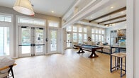 This single-floor community clubhouse offers a generous, open-plan layout designed for both relaxation and socializing. The entrance welcomes visitors with soaring ceilings, large transom-topped French doors, and light hardwood flooring that flows seamlessly throughout the space. A bold teal accent wall adorned with curated artwork and a built-in banquette adds a vibrant focal point near the entry.

The expansive living room is the heart of the space, anchored by exposed dark wood ceiling beams, shiplap detailing, and an elegant chandelier. Floor-to-ceiling windows along the perimeter flood the interior with natural light while offering views of the surrounding outdoor landscape. A full-size pool table serves as a central recreational feature, complemented by multiple seating arrangements featuring neutral sofas and accent chairs arranged on patterned area rugs.

A dedicated workstation area with bar-height desks and computer terminals provides a functional workspace, while a kitchenette with full-size refrigerator, gray cabinetry, and a geometric tile backsplash supports community gatherings. A live-edge wood communal dining table surrounded by upholstered chairs adds warmth and character. Throughout, the design balances coastal-inspired tones with modern furnishings, creating a welcoming and well-appointed communal environment.