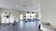 This fitness center is a clean, single-floor facility designed to support a well-rounded workout routine. The space features a row of treadmills and elliptical machines aligned along the windowed wall, allowing users to exercise with the benefit of natural light streaming in through multiple windows. The dark, rubberized flooring provides both durability and safety, making it well-suited for a variety of exercises. Strength training is accommodated with a weight bench and additional equipment positioned along the side wall. Multiple wall-mounted flat-screen televisions are installed at convenient viewing angles, enhancing the cardio experience. Recessed lighting in the white ceiling ensures the space remains bright and well-illuminated at all times. The neutral, light-gray walls contribute to a clean and open atmosphere throughout the room. A water fountain is conveniently located near the entrance, providing easy access for hydration. A door with a window offers direct access to the exterior, adding to the overall sense of openness. The layout is thoughtfully organized to maximize usable floor space, allowing for comfortable movement between equipment stations.