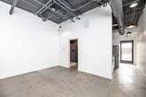Located in Des Moines, Iowa, this single-floor unit showcases a distinctive industrial aesthetic throughout its efficiently designed layout. Polished concrete floors run continuously across the space, complementing the bold exposed ceiling system that features visible ductwork, conduit, and structural elements finished in dark tones — a hallmark of urban, loft-inspired design.

The main living area is open and versatile, wrapped in crisp white walls that contrast the raw industrial ceiling overhead. Pendant lighting with warm-toned fixtures provides focused illumination, enhancing the clean, minimalist atmosphere. A kitchen area is equipped with stainless steel appliances, including a full-size refrigerator and range, tucked efficiently along one wall.

The bathroom offers a thoughtfully finished retreat, featuring large-format dark tile in the walk-in shower with matte black fixtures, a standard toilet, and polished concrete floors that continue from the main living space. An adjacent closet provides convenient built-in storage with a hanging rod.

An electrical panel is visibly mounted on the white concrete wall, consistent with the space's industrial character. The laundry area adds practical functionality. Throughout, the combination of white walls, raw concrete, and exposed mechanical systems creates a cohesive, modern industrial ambiance well-suited for a variety of uses.