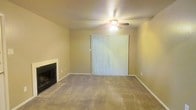 This single-floor residential space offers a comfortable and functional layout designed for everyday living. The living room serves as the heart of the home, anchored by a classic fireplace with a tiled hearth that provides a warm and inviting focal point. Soft, wall-to-wall carpeting extends throughout the space, adding a sense of warmth underfoot. A ceiling fan with an integrated light fixture is centrally positioned overhead, offering both air circulation and ambient lighting. A large sliding glass door, dressed with vertical blinds, fills the room with natural light and provides direct access to the outdoors, creating a seamless connection between the interior and exterior. The neutral, warm-toned walls offer a versatile backdrop, easily adaptable to a variety of furnishing styles and décor preferences. The overall ambiance is calm and understated, making it a practical and livable space well-suited for those seeking a straightforward, single-level home environment.
