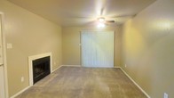 This single-floor residential space offers a comfortable and functional layout designed for everyday living. The living room serves as the heart of the home, anchored by a classic fireplace with a tiled hearth that provides a warm and inviting focal point. Soft, wall-to-wall carpeting extends throughout the space, adding a sense of warmth underfoot. A ceiling fan with an integrated light fixture is centrally positioned overhead, offering both air circulation and ambient lighting. A large sliding glass door, dressed with vertical blinds, fills the room with natural light and provides direct access to the outdoors, creating a seamless connection between the interior and exterior. The neutral, warm-toned walls offer a versatile backdrop, easily adaptable to a variety of furnishing styles and décor preferences. The overall ambiance is calm and understated, making it a practical and livable space well-suited for those seeking a straightforward, single-level home environment.