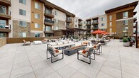 Located in Draper, Utah, this expansive rooftop terrace offers a well-appointed outdoor amenity space with stunning panoramic views of the surrounding mountain ranges. The large-format porcelain tile deck provides a clean, modern foundation throughout the entire level, creating a cohesive and polished aesthetic.

The terrace is thoughtfully divided into multiple functional zones. Generous lounge seating areas feature a mix of wicker sectional sofas, sleek modular ottomans, and cushioned arm chairs accented with vibrant orange throw pillows, centered around fire pit tables ideal for evening gatherings. Sun loungers with bold umbrella shading offer dedicated relaxation areas for daytime use.

A pergola structure anchors one section of the rooftop, strung with bistro lighting and framing an outdoor dining area complete with long communal tables and chairs. Adjacent to this covered zone is a BBQ station equipped with grills, providing a fully functional outdoor cooking and entertaining space. A hot tub is also integrated into the layout, adding a resort-like amenity to the rooftop experience.

The perimeter of the terrace features low-profile walls and open railings that preserve sightlines to the dramatic Utah mountain backdrop and expansive sky. The surrounding residential building showcases a contemporary design with stone and wood accents, balconies, and large windows that overlook the shared rooftop space below.