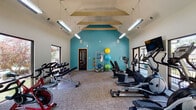 Located at 201 Heritage Hill Way in Louisville, Kentucky, this well-appointed fitness center is designed to support a comprehensive workout experience. The space is defined by its striking vaulted ceiling with exposed decorative beams and a bold teal accent wall that energizes the interior atmosphere. Suspended fluorescent lighting runs the length of the room, ensuring the space is brightly and evenly illuminated throughout.

Large windows line multiple walls, flooding the room with natural light and offering views of the surrounding greenery, creating an open and inviting environment. The fitness center is equipped with a wide variety of exercise machines, including treadmills, elliptical trainers, stair climbers, and stationary cycling bikes. Wall-mounted flat-screen televisions are positioned near the cardio equipment for entertainment during workouts.

A dedicated free weights area features a dumbbell rack alongside a weight bench, catering to strength training needs. Additional functional fitness equipment is neatly organized along the teal accent wall, including stability balls in multiple sizes and resistance training accessories. The flooring throughout consists of durable, low-pile carpet tile suited for high-traffic fitness use. A secondary door provides direct outdoor access, further enhancing convenience and ventilation within this thoughtfully designed community fitness facility.