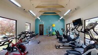 Located at 201 Heritage Hill Way in Louisville, Kentucky, this well-appointed fitness center is designed to support a comprehensive workout experience. The space is defined by its striking vaulted ceiling with exposed decorative beams and a bold teal accent wall that energizes the interior atmosphere. Suspended fluorescent lighting runs the length of the room, ensuring the space is brightly and evenly illuminated throughout.

Large windows line multiple walls, flooding the room with natural light and offering views of the surrounding greenery, creating an open and inviting environment. The fitness center is equipped with a wide variety of exercise machines, including treadmills, elliptical trainers, stair climbers, and stationary cycling bikes. Wall-mounted flat-screen televisions are positioned near the cardio equipment for entertainment during workouts.

A dedicated free weights area features a dumbbell rack alongside a weight bench, catering to strength training needs. Additional functional fitness equipment is neatly organized along the teal accent wall, including stability balls in multiple sizes and resistance training accessories. The flooring throughout consists of durable, low-pile carpet tile suited for high-traffic fitness use. A secondary door provides direct outdoor access, further enhancing convenience and ventilation within this thoughtfully designed community fitness facility.