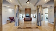 This single-story community facility makes a striking first impression through a glass-paneled entry flanked by tall, reclaimed wood-tiled columns and an arched ceiling with an industrial-style pendant light. The grand lobby flows seamlessly into a spacious lounge area anchored by a soaring floor-to-ceiling wood-clad fireplace wall, mounted with a flat-screen television. Comfortable mid-century modern seating — including leather sofas and sculptural wood-framed chairs — is arranged atop a colorful area rug, creating an inviting communal gathering space. Recessed lighting and vaulted ceilings amplify the sense of openness throughout. Adjacent to the lounge, a casual dining area features a rustic wood communal table with eclectic seating, illuminated by modern pendant lights. Floor-to-ceiling sliding glass doors open directly to an outdoor patio shaded by mature palm trees, flooding the interior with natural light and creating a strong indoor-outdoor connection. The fitness center is well-equipped with a comprehensive selection of cardio machines, weight equipment, and mirrors, enlivened by a bold geometric mural. Throughout the space, large-format tile flooring with inlaid wood-tone accents ties each area together cohesively, conveying a modern yet warm aesthetic.