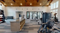 This expansive fitness center offers a thoughtfully designed workout environment spanning a generous single-floor layout. The space is immediately distinguished by its dramatic vaulted ceiling clad in rich wood planking, supported by exposed beams and complemented by large ceiling fans that keep the air circulating throughout. Recessed and pendant lighting illuminates the interior, while an accent wall of exposed brick adds warmth and character to the overall aesthetic.

Natural light pours in through floor-to-ceiling windows and glass-panel doors on multiple sides of the facility, offering outdoor views and creating an open, energizing atmosphere. The main workout area is carpeted in a multi-toned striped pattern and is organized to accommodate a wide variety of fitness activities. Cardio equipment — including treadmills, ellipticals, and stationary cycling bikes with integrated screens — lines the perimeter near the windows.

A dedicated strength training zone features Matrix cable and multi-use machines, free-weight racks, a rowing machine, and an adjustable bench. A functional training area is equipped with a power rack, TRX suspension straps, a punching bag, medicine balls, and stability balls. A central storage island provides convenient access to fitness accessories. A separate bright annex room with large windows and light wood-style flooring houses additional cycling stations, offering a semi-private spin area with outdoor views.
