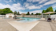 Located in West Jordan, Utah, this outdoor community pool facility offers a well-appointed recreational space designed for relaxation and leisure. The centerpiece is a generously sized rectangular swimming pool with clear, blue-tinted water, surrounded by an expansive concrete pool deck that provides ample room for lounging and socializing.

The deck is furnished with multiple reclining lounge chairs positioned along the perimeter of the pool, offering convenient spots to relax in the sun. A standout architectural feature is a white pergola structure positioned on one side of the deck, providing a shaded seating area complete with bistro-style table and chairs—ideal for casual gatherings. A second pergola is also visible on the opposite end, further enhancing the amenity space.

The entire area is enclosed by a white vinyl fence, ensuring privacy and safety while maintaining an open, airy feel. Mature trees lining the exterior of the fence contribute natural greenery and additional shade. A small utility building and what appears to be a vending machine are accessible from the deck, adding everyday convenience to the space. The surrounding neighborhood rooftops and lush tree canopy are visible beyond the fence, situating this facility within an established residential community.