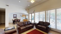 This single-floor space is designed to foster a comfortable, communal atmosphere through a well-organized layout that seamlessly connects several functional areas. The entrance greets visitors with a bright, tiled foyer framed by double glass doors that flood the space with natural light and provide views of the surrounding exterior.

The main lounge area is generously proportioned, featuring soaring ceilings, warm cream-toned walls, and large sliding glass doors that open to outdoor views, likely a pool area. Dark leather sofas and armchairs are arranged around a red area rug, while a sleek fireplace anchors one wall alongside wall-mounted artwork. An arched alcove houses a flat-screen television above a black media console, adding a dedicated entertainment corner to the space.

The kitchen is fully equipped with honey-toned cabinetry, a gas range, microwave, refrigerator, dishwasher, and a double sink beneath a window that brings in additional natural light. Light ceramic tile flooring runs consistently throughout the space, reinforcing the cohesive design.

A separate dining area accommodates a raised wooden table surrounded by upholstered high-back chairs, ideal for group dining. Framed artwork throughout adds character and warmth to the overall environment.