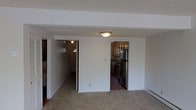 This two-floor residential space offers a practical and comfortable layout designed for everyday living. The main level centers around a carpeted living area that flows openly toward two distinct doorways — one leading to the kitchen and another to the entryway and staircase. The kitchen is well-equipped with dark wood cabinetry, a stainless steel refrigerator, and a matching range, all set on hardwood-style flooring that contrasts with the soft carpeting of the living area. Flush-mount ceiling lighting provides consistent illumination throughout the main level. The entryway area features a front door with a transom window that allows natural light to filter in, and a staircase that connects the main floor to the upper level. A baseboard heating unit is visible along the living area wall, indicating a reliable in-unit heating system. The overall aesthetic is clean and straightforward, with neutral wall tones and a layout that emphasizes functionality. The second floor provides additional living space above, completing a multi-level floor plan suited for a range of occupants.