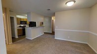 This single-floor residence offers a practical and open layout that integrates the kitchen and living areas into one cohesive space. The kitchen is equipped with black appliances, including a refrigerator, microwave, and range, complemented by warm honey-toned cabinetry and granite-style countertops. A peninsula breakfast bar serves as a natural divider between the kitchen and the adjoining living area, providing both additional counter space and a casual dining option. The living area is finished with neutral-toned carpeting that adds warmth underfoot, while the walls are painted in a soft, neutral tone that enhances the sense of openness. A textured ceiling runs throughout the space, and flush-mount lighting provides consistent illumination. HVAC vents are integrated into the ceiling for climate control. A hallway visible from the main living area leads to additional rooms, suggesting a well-organized floor plan. White baseboards and trim provide clean contrast against the neutral walls, contributing to a tidy, well-maintained aesthetic throughout the home.