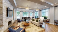 Located at 510 N Wilmington St in Raleigh, NC, this elegantly designed single-floor lounge space offers a refined and welcoming atmosphere. The open-concept layout is unified by striking herringbone-patterned hardwood flooring that flows seamlessly throughout the entire space.

The focal point of the main lounge area is a dramatic floor-to-ceiling stacked stone accent wall, complete with a sleek linear fireplace, a wall-mounted television, and flanking decorative niches with built-in lighting. This feature wall anchors a well-appointed seating area furnished with a cream sofa, mid-century modern accent chairs, and an organic-shaped wood coffee table atop a gold-toned area rug.

Expansive floor-to-ceiling windows line multiple walls, flooding the space with abundant natural light and offering clear views of the lush greenery outside. Sheer white drapery panels frame the windows, adding an airy, sophisticated touch. Coffered ceilings with recessed lighting and large pendant drum fixtures contribute to the elevated architectural character of the space.

A high-top glass and wood bar table with upholstered counter chairs provides a dedicated seating area near the windows, ideal for collaborative use. The kitchenette area is outfitted with dark cabinetry, white countertops, stainless steel appliances including a refrigerator and microwave, and a decorative mosaic tile backsplash. A separate seating alcove with lounge chairs near additional windows completes this versatile and polished space.