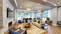 Located at 510 N Wilmington St in Raleigh, NC, this elegantly designed single-floor lounge space offers a refined and welcoming atmosphere. The open-concept layout is unified by striking herringbone-patterned hardwood flooring that flows seamlessly throughout the entire space.

The focal point of the main lounge area is a dramatic floor-to-ceiling stacked stone accent wall, complete with a sleek linear fireplace, a wall-mounted television, and flanking decorative niches with built-in lighting. This feature wall anchors a well-appointed seating area furnished with a cream sofa, mid-century modern accent chairs, and an organic-shaped wood coffee table atop a gold-toned area rug.

Expansive floor-to-ceiling windows line multiple walls, flooding the space with abundant natural light and offering clear views of the lush greenery outside. Sheer white drapery panels frame the windows, adding an airy, sophisticated touch. Coffered ceilings with recessed lighting and large pendant drum fixtures contribute to the elevated architectural character of the space.

A high-top glass and wood bar table with upholstered counter chairs provides a dedicated seating area near the windows, ideal for collaborative use. The kitchenette area is outfitted with dark cabinetry, white countertops, stainless steel appliances including a refrigerator and microwave, and a decorative mosaic tile backsplash. A separate seating alcove with lounge chairs near additional windows completes this versatile and polished space.