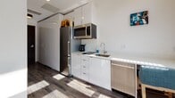 This single-floor studio is thoughtfully designed to maximize functionality within a compact, open layout. The main living area seamlessly integrates sleeping and lounge spaces, featuring light wood-style flooring that runs throughout and contributes to a clean, contemporary aesthetic. A standout design element is the bold herringbone-patterned feature wall in deep blue-grey tones, which anchors the sleeping area and adds visual depth to the otherwise neutral white surroundings.

Large windows along one wall allow generous natural light to flood the space, offering views of the surrounding streetscape and creating a bright, airy atmosphere. The sleeping area accommodates a full bed with a wood-frame base, while a grey sectional sofa defines the lounge corner, complemented by colorful wall art that adds personality to the clean palette.

The compact kitchenette is efficiently outfitted with high-gloss white cabinetry, stainless steel appliances including a refrigerator, microwave, dishwasher, and an induction cooktop with an integrated sink, providing all essential amenities in a streamlined design.

The bathroom features large-format tiles in warm taupe and dark charcoal tones on both the floors and walls, lending a polished, modern finish. A wall-mounted vanity and standard toilet complete the well-appointed space.