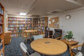 This single-floor library space is thoughtfully designed to accommodate avid readers and casual browsers alike. The room is lined on nearly every wall with tall, light wood bookshelves packed from top to bottom with an extensive and diverse collection of books spanning numerous genres. The sheer volume of titles on display gives the space a rich, well-curated character.

Two distinct seating areas serve different needs within the room. A round wooden table with lightweight chairs occupies the central area, illuminated by a skylight overhead that introduces natural light into the space. A separate, more relaxed lounge area features cushioned armchairs arranged around a small side table, offering a comfortable spot for extended reading sessions. Decorative touches such as framed artwork, a small potted plant, and a table lamp contribute warmth to the otherwise functional layout.

The flooring throughout is a blue-toned carpet that softens the room acoustically and visually. A dark wood display cabinet near the entrance showcases featured titles, while a corkboard on the wall provides space for notices. The drop ceiling with recessed and overhead lighting ensures the room remains well-lit throughout. The overall ambiance is calm, organized, and inviting—well-suited for quiet reading and literary exploration.