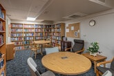 This single-floor library space is thoughtfully designed to accommodate avid readers and casual browsers alike. The room is lined on nearly every wall with tall, light wood bookshelves packed from top to bottom with an extensive and diverse collection of books spanning numerous genres. The sheer volume of titles on display gives the space a rich, well-curated character.

Two distinct seating areas serve different needs within the room. A round wooden table with lightweight chairs occupies the central area, illuminated by a skylight overhead that introduces natural light into the space. A separate, more relaxed lounge area features cushioned armchairs arranged around a small side table, offering a comfortable spot for extended reading sessions. Decorative touches such as framed artwork, a small potted plant, and a table lamp contribute warmth to the otherwise functional layout.

The flooring throughout is a blue-toned carpet that softens the room acoustically and visually. A dark wood display cabinet near the entrance showcases featured titles, while a corkboard on the wall provides space for notices. The drop ceiling with recessed and overhead lighting ensures the room remains well-lit throughout. The overall ambiance is calm, organized, and inviting—well-suited for quiet reading and literary exploration.