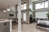 This two-story loft offers a striking industrial-modern aesthetic defined by soaring double-height ceilings and exposed silver ductwork that runs throughout the main living level. Light wood-tone flooring flows continuously across both floors, complementing the neutral gray and beige wall palette and creating a cohesive, contemporary feel throughout.

The open-concept main level integrates the living, dining, and kitchen areas into one expansive, light-filled space. Large windows fitted with horizontal blinds admit generous natural light, while the upper-level clerestory windows add additional brightness and visual height. Structural columns punctuate the floor plan, reinforcing the loft's architectural character.

The kitchen is well-appointed with wood-grain cabinetry, stainless steel appliances including a French-door refrigerator, a white tile backsplash, and light quartz countertops. A peninsula island with seating for multiple occupants is illuminated by pendant lighting, serving as both a functional workspace and a casual dining area.

The bedroom features the same dramatic ceiling height with exposed ductwork and a ceiling fan, alongside multiple windows that provide ample natural light. A full bathroom with a walk-in shower and wood-tone vanity serves the bedroom, while a separate half bath is located on the main level.