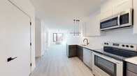 This thoughtfully designed single-floor unit offers a clean, contemporary aesthetic defined by bright white walls, light-toned luxury vinyl plank flooring, and bold black accent hardware throughout. The open-concept kitchen flows seamlessly into the living area, creating a connected and functional layout. The kitchen is outfitted with a full suite of stainless steel appliances—including a refrigerator, electric range, built-in microwave, and dishwasher—complemented by two-tone cabinetry featuring crisp white upper cabinets and dark lower cabinets, topped with light quartz countertops. A modern pendant light fixture adds a stylish focal point above the kitchen peninsula, which includes a single-basin sink with a matte black faucet. The living area is open and airy, offering a generous amount of natural light. Multiple bedrooms provide comfortable, well-proportioned spaces, each with large black-framed windows that frame views of the surrounding neighborhood and allow abundant daylight to fill the rooms. Each bedroom also features a wall-mounted HVAC unit for individual climate control. The bathroom is finished with a walk-in shower enclosed by a frameless glass panel, featuring a matte black rainfall showerhead and matching fixtures against crisp white surround walls, delivering a clean, spa-inspired feel.