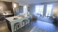 This thoughtfully designed single-floor space showcases a seamless open-concept layout that connects the kitchen, dining, and living areas into one cohesive environment. The kitchen is outfitted with stainless steel appliances, including a French door refrigerator with an ice and water dispenser, and features a wide white countertop island with an undermount sink. The island doubles as a casual breakfast bar, accommodating multiple industrial-style metal stools that add a contemporary edge to the space. Pendant lighting above the island provides focused illumination and a stylish visual anchor. A chalkboard accent wall behind the kitchen adds a playful, functional touch. The dining area sits just beyond the kitchen, featuring a glass-top table with modern white chairs. The living room is bathed in natural light streaming through expansive floor-to-ceiling windows that open to an outdoor area, offering views of lush greenery. Rich wood-tone flooring runs throughout, grounding the space with warmth, while an area rug beneath the living space adds texture and definition. The overall ambiance is clean, modern, and inviting, blending functional design with contemporary aesthetics.