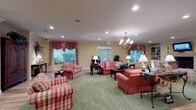 Located at 1955 Poppy Ferry Rd in Biloxi, Mississippi, this single-floor facility exudes a welcoming, Southern-inspired ambiance throughout its thoughtfully designed common areas. The main lounge is a generously proportioned gathering space adorned with soft green carpeting, warm yellow walls, and crown molding. Multiple seating arrangements feature coral and plaid upholstered sofas and armchairs centered around ornate wooden coffee tables, creating intimate conversation areas. An elegant chandelier anchors the room overhead, while recessed lighting provides even illumination throughout. Large windows dressed with decorative red and pink valances and French doors flood the space with natural light and offer views of the lush outdoor greenery surrounding the property. A secondary lounge area off the main space includes a classic white fireplace mantel topped with a wall-mounted television, built-in cabinetry, and hardwood-style flooring, adding a cozy, residential feel. A dedicated refreshment station features a wraparound counter with a light stone surface, white cabinetry, and a beverage service setup, providing a convenient amenity for occupants. Throughout the facility, the consistent use of warm tones, traditional furnishings, ornamental accents, and abundant natural light creates a comfortable, homelike environment that is both functional and inviting.
