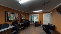 Located in Conroe, TX, this single-floor business center is designed to support productivity and convenience in a welcoming environment. The space is defined by warm, earthy orange walls accented with crisp white crown molding, giving the interior a vibrant yet professional character. Patterned carpet flooring ties the space together throughout.

The primary workstation area features a built-in granite-top counter running along one wall, lined with multiple desktop computers and ergonomic task chairs, providing ample workspace for individuals needing computing resources. Two large framed abstract art pieces in bold, multicolored tones add a creative touch to the workstation wall.

Natural light filters in through large windows fitted with horizontal blinds, offering views of the surrounding greenery outside. A small round bistro-style table with accompanying chairs is positioned near the window, serving as a casual spot for meetings or quiet work.

A dedicated lounge area includes black leather armchairs arranged around a low side table, with a wall-mounted flat-screen television positioned for easy viewing. Overhead fluorescent lighting ensures the entire space remains well-lit throughout the day. Interior doorways connect to adjacent areas, enhancing the overall flow and functionality of this practical, amenity-focused business center.