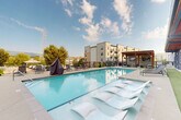 Located at 726 Fulton St in Chattanooga, Tennessee, this outdoor amenity space centers around a generously sized, resort-style swimming pool with crystal-clear blue water. The pool deck is outfitted with a mix of modern white in-water lounge chairs and traditional gray chaise lounges, providing ample space for sunbathing and relaxation. A covered pavilion with warm wood-beam detailing anchors one side of the deck, offering a shaded retreat with ambient lighting for evening use. Large glass folding doors connect the pool area directly to an indoor lounge or clubroom, seamlessly blending indoor and outdoor living. The surrounding deck is finished in a clean, light-toned concrete, bordered by black metal fencing that frames sweeping views of Chattanooga's rolling green hillside. A covered outdoor grilling station is also available on the deck. The multi-story residential building rises as a striking backdrop, featuring contemporary architecture with large-paned windows and private balconies. The overall ambiance is open, airy, and modern, capturing the natural beauty of the surrounding Tennessee landscape while delivering a polished, community-focused outdoor experience.