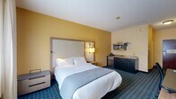 Located at 1383 Washington Ave in Albany, NY, this efficiently designed guest room offers a comfortable and functional layout across a single floor. The spacious main bedroom is the focal point of the unit, featuring a queen-sized bed dressed in crisp white linens and anchored by a contemporary upholstered headboard with integrated wall sconces on either side. Warm golden-yellow accent walls add a welcoming vibrancy to the room, while a boldly patterned teal and blue carpet grounds the space with visual interest.

Natural light filters in through sheer-curtained windows, complementing the room's bright, neutral ceiling and walls. A dedicated work area includes a full-sized desk, ergonomic chair, and a wall-mounted flat-screen television above, making the room well-suited for both leisure and business travelers. A compact kitchenette along one wall provides a mini-refrigerator, microwave, and sink with overhead shelving for added convenience.

The bathroom features large-format light tile flooring, a floating wood-toned vanity with a gray countertop and integrated sink, a wide mirror with overhead lighting, and a striking deep navy herringbone-patterned accent wall that adds a modern, designer touch. A hallway connects the main living area to the bathroom, completing this thoughtfully arranged room.