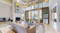 Located at 13100 W Bellfort Ave in Houston, Texas, this single-floor community clubhouse is designed to impress with its refined architectural details and welcoming atmosphere. The entrance foyer sets a grand tone immediately, featuring soaring double-height ceilings, light hardwood flooring, a central round pedestal table, and an abundance of natural light pouring in through tall windows and glass-paneled French doors. Elegant chandelier lighting adds warmth and sophistication throughout.

The hallway connects the foyer to the main lounge area, which is anchored by a classic white-trimmed fireplace and flanked by floor-to-ceiling windows dressed in bold ikat-patterned drapery. Views of the outdoor pool area are visible through the expansive glazing. The lounge is furnished with plush neutral sofas, a sleek coffee table, and accented with yellow and white decorative pillows, creating an inviting and polished communal space.

Arched architectural niches, built-in shelving, tray ceilings with recessed lighting, and crown molding further elevate the interior's character. The warm beige and taupe color palette is consistent throughout, lending a cohesive and upscale feel. A secondary lounge area with access to a dining or business space via French doors adds functional versatility to this well-appointed community gathering space.