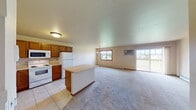 This single-floor unit is thoughtfully laid out with an open-concept design that seamlessly connects the kitchen and living area, creating a spacious, versatile environment. The kitchen is well-equipped with warm oak cabinetry extending to the ceiling for generous storage, light-toned countertops, a double stainless steel sink, and white appliances including an electric range, over-the-range microwave, and refrigerator. Tile flooring runs throughout the kitchen, while a central island provides additional prep and seating space. The adjoining living area is expansive and finished with neutral carpet, baseboard heating, and a wall-mounted air conditioning unit. A sliding glass door opens to a private balcony, allowing natural light to fill the room. A pendant chandelier adds a subtle decorative touch to the dining area. The bathroom features tile flooring, an oak vanity with a white countertop, a toilet, towel bars, and a shower-tub combination. Throughout the unit, neutral tones and oak wood accents create a cohesive, understated aesthetic suited for everyday living.
