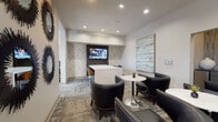 Located at 8718 Wintersweet Ln in Charlotte, NC, this single-floor community business center and lounge offers a well-appointed space designed for both productivity and relaxed gatherings. The interior features a contemporary aesthetic defined by neutral gray tones, textured herringbone accent walls, and patterned carpeting that adds visual depth throughout.

The lounge area serves as the heart of the space, furnished with sleek, studded black leather club chairs arranged around round marble-top pedestal tables. This seating arrangement encourages conversation and informal meetings, while a wall-mounted flat-screen television provides entertainment. Carefully curated artwork, including a large abstract canvas in soft blues and greens, adds a refined, gallery-like quality to the walls alongside bold, sculptural sunburst mirror groupings.

A dedicated workstation area is equipped with a computer, printer, and ample desk space, supported by natural light streaming through large windows fitted with horizontal blinds. A warm wood-toned credenza provides additional storage, complementing the room's sophisticated palette.

The space benefits from recessed ceiling lighting throughout, ensuring a bright and welcoming ambiance at all times. Glass-paneled entry doors connect the lounge to adjacent areas, reinforcing an open, accessible feel. Overall, this business center combines functional design with upscale finishes to create a versatile environment suited for work, meetings, or leisure.