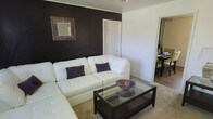 This single-floor space showcases a thoughtfully designed interior that balances bold design choices with a clean, modern aesthetic. The living area serves as the centerpiece, featuring a striking black accent wall that creates a dramatic focal point against the surrounding white walls. A decorative sunburst-style mirror adorns the accent wall, adding visual interest and depth to the space. The room is furnished with a plush white tufted sectional sofa accented with deep purple throw pillows, creating a sophisticated contrast of light and dark tones. A dark-framed glass coffee table sits atop a soft area rug, anchoring the seating arrangement and providing a functional yet elegant surface. The flooring throughout features warm hardwood tones, adding a sense of continuity and warmth to the space. An open passageway connects the living area to the dining space, which is visible in the background and features a dark dining set, reinforcing the cohesive dark-and-light design theme carried throughout the home. The overall ambiance of the space is contemporary and polished, with a carefully curated color palette of black, white, and purple that lends the interior a refined, stylish character.
