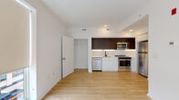 This contemporary two-floor space showcases a clean, modern design with light hardwood flooring running throughout both levels, creating a cohesive and airy aesthetic. The open-concept lower level seamlessly connects the living area to the kitchen, which serves as a functional centerpiece of the home. The kitchen is outfitted with a striking two-tone cabinet design, pairing dark espresso upper cabinets with crisp white lower cabinetry. A full suite of stainless steel appliances—including a refrigerator, electric range, built-in microwave, and dishwasher—complement the white tile backsplash and light countertops. Under-cabinet lighting adds a refined touch, while a matte black faucet over a single-basin sink introduces a subtle contrast. Recessed lighting illuminates both floors throughout. A staircase connects to the upper level, where a bright, open room benefits from large floor-to-ceiling windows fitted with roller shades, inviting generous natural light. The bathroom features large-format travertine-style floor and wall tiles surrounding a soaking tub with a showerhead, accented by dark oil-rubbed bronze fixtures for an elevated, spa-like feel. The overall design is minimal and contemporary.
