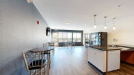 This single-floor dining and kitchen space offers a practical and well-appointed layout designed for communal use. The open floor plan is anchored by durable wood-look flooring that flows seamlessly throughout the entire area, complementing the cool blue-gray accent walls that define the room's contemporary aesthetic.

The dining area accommodates multiple high-top tables paired with cushioned bar-height stools, creating a casual and social atmosphere. A wall-mounted flat-screen television is positioned for easy viewing from the seating area. Along the far wall, a generous row of large windows fitted with roller shades floods the space with natural light and provides views of an outdoor balcony beyond. A glass door leads directly outside, enhancing the connection between interior and exterior.

The kitchen section features warm-toned wood cabinetry with dark granite countertops, a stainless steel sink, and a peninsula counter that serves as both a prep surface and a functional divider. Pendant lighting above the kitchen area adds a polished touch. Upper cabinets provide ample storage, and a microwave and refrigerator complete the kitchen's practical amenities. A vending or display unit near the kitchen entrance adds additional convenience to this well-organized communal space.