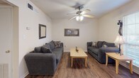 This single-story residence offers a thoughtfully organized layout that balances comfort and practicality throughout its key living spaces. The living room serves as a central gathering area, featuring rich hardwood-style flooring, a ceiling fan with light fixture, and neutral wall tones that create a calm, welcoming atmosphere. Two sofas and a central coffee table define the space efficiently.

Adjacent to the living room, the dining area continues the hardwood-style flooring and is illuminated by a pendant chandelier overhead, providing a well-lit setting suitable for everyday meals or entertaining. The space connects conveniently to the front entry.

The kitchen features a galley-style configuration with ample cabinetry in a light gray finish, speckled countertops, a double sink, and black appliances including an electric range. The hardwood-style flooring carries through here as well, enhancing visual continuity.

The bedroom is carpeted for comfort and includes a ceiling fan, a window allowing natural light, and sufficient space for a full furniture arrangement. A hallway area with additional storage connects the bedroom to the bathroom, which includes a wide vanity with speckled countertop and a full tub-shower combination.