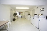 This single-floor shared laundry facility is designed with practicality and functionality in mind. The space is finished with light-colored walls and speckled terrazzo-style flooring, creating a clean and well-maintained environment. Fluorescent lighting runs along the textured ceiling, ensuring the entire area is brightly and evenly illuminated.

The laundry area is equipped with multiple top-loading washing machines and dryers, each featuring card-operated payment panels for convenient, cashless use. Machines are arranged along the walls, allowing for comfortable circulation throughout the space. A utility sink is also present, providing additional functionality for residents.

A central folding and sorting area includes a standard folding table paired with a freestanding garment rack, offering practical space for managing laundry. Seating is available nearby with a plastic chair, making wait times more comfortable. A decorative potted plant and a colorful hot air balloon artwork add a welcoming touch to the otherwise utilitarian space.

Safety features are clearly in place, including a mounted fire extinguisher and illuminated exit signage. A small skylight allows a modest amount of natural light to supplement the overhead fixtures. Overall, the facility is straightforward, clean, and well-equipped for shared residential use.