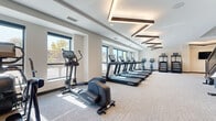 This contemporary fitness facility spans two floors and is thoughtfully designed to support a comprehensive workout experience. The main cardio area is bright and spacious, lined with large windows that invite ample natural light and offer views of the surrounding outdoor environment. A row of treadmills and elliptical machines from Life Fitness is arranged along the windowed walls, creating an open and energizing atmosphere. Geometric, rectangular pendant lighting fixtures are suspended from coffered ceilings, adding a modern architectural detail throughout the space.

A dedicated free weights zone features multi-tier dumbbell racks, a weight bench, and floor-to-ceiling mirrors that enhance the sense of depth and space. Additional strength training machines and functional training equipment, including cable systems and resistance machines, are distributed across both levels. A corridor connecting the workout areas is accented by a bold, colorful geometric mural that runs along the staircase, serving as a striking visual focal point.

The second floor offers additional training space with wall-mounted televisions, stability equipment, and a punching bag. A standout amenity is the private sauna room, fully lined in natural cedar wood with tiered benches and a traditional electric heater, providing a tranquil space for recovery and relaxation.